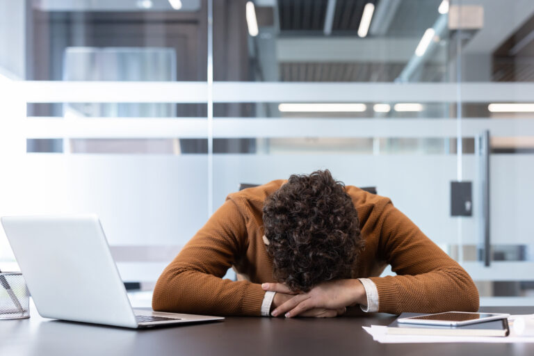 A sad person rests his head on their arms atop a desk, thinking about his career success.